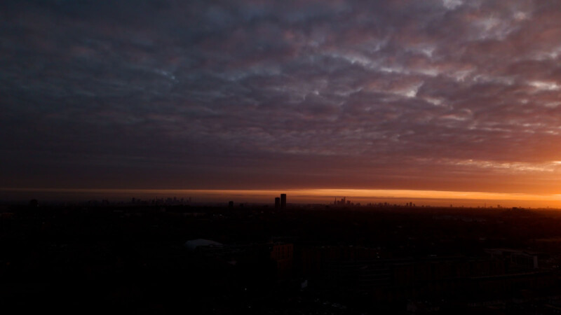 A city skyline is silhouetted against a dramatic sunset. Dark clouds blanket the sky, with a strip of orange and yellow light near the horizon, casting a warm glow over the scene.