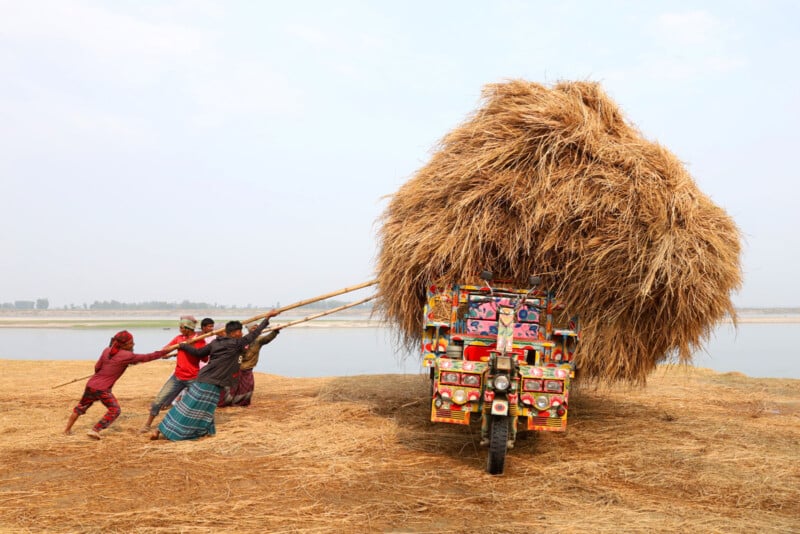 A group of people push a colorful, overloaded vehicle stacked with hay near a river. The hay is piled high, almost tipping. The scene takes place on a sandy, straw-covered riverbank under a clear sky.