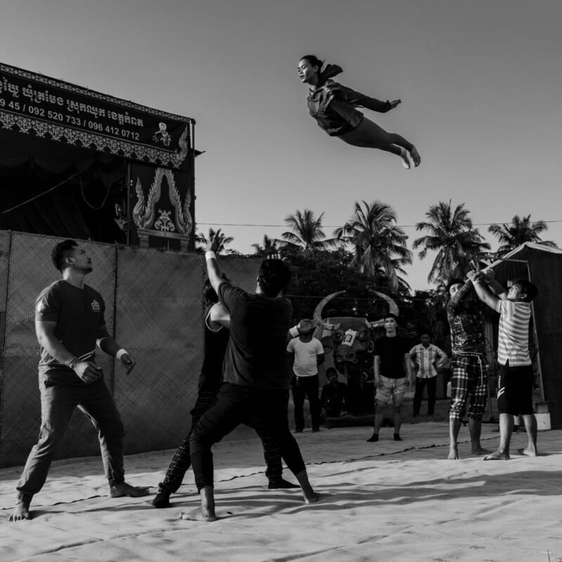 A black and white image of a group of people performing an acrobatic stunt outdoors. Several individuals on the ground support a person mid-air, arms extended. Palm trees and people watching are in the background.