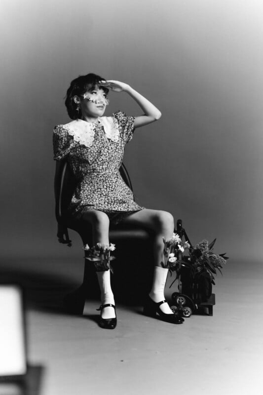 A person in a floral dress with a white collar sits on a chair, saluting. They wear socks and black shoes. Plants are placed in a cart near the chair. The photo is in black and white, with a studio background.