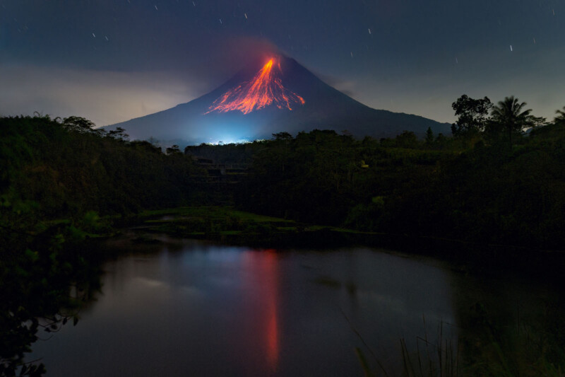 A nighttime scene of an erupting volcano with lava glowing at its peak. The bright orange and red lava contrasts against the dark sky, while a calm lake in the foreground reflects the fiery colors. Dense vegetation surrounds the area.