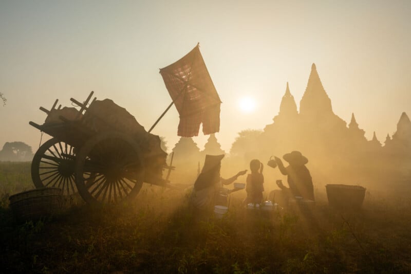 Silhouetted against a sunset, a family sits on the grass near a cart, enjoying a meal. Ancient pagodas are visible in the misty background, creating a serene and timeless scene.