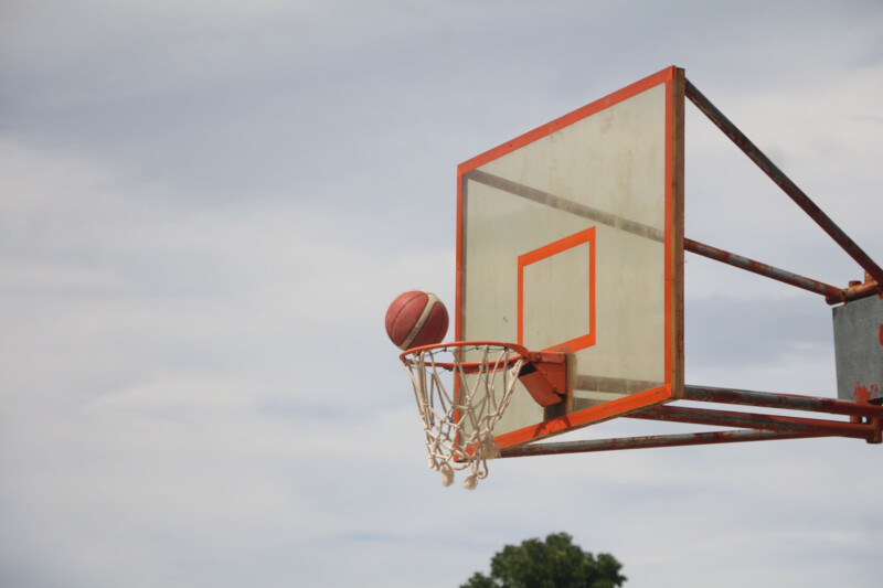 A basketball is passing through a red and white hoop, with a clear, cloud-dotted sky in the background. A green treetop is visible at the bottom of the frame.