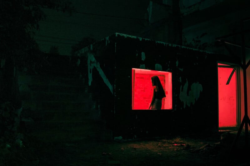 A dimly lit scene shows a dark building with two rectangular, red-lit windows. A silhouette of a person is visible through the larger window. A stairway leads up into darkness on the left. The overall atmosphere feels mysterious and eerie.