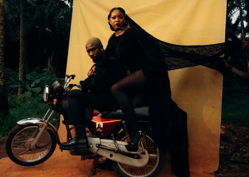 A man and woman in black clothing pose on a motorcycle. The woman wears a long lace veil held up by an unseen person. A yellow backdrop and tropical plants form the background, adding contrast to the scene.