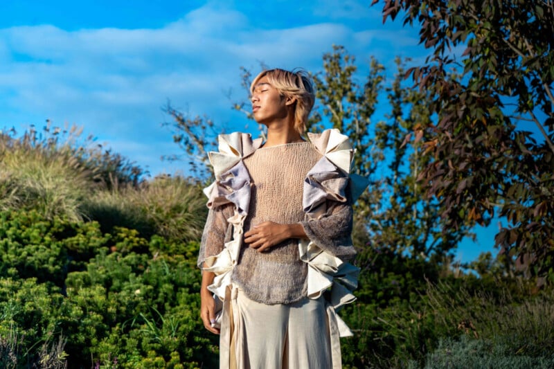 A person with light hair stands outdoors, eyes closed, wearing a unique beige outfit with large, ornate ruffles on the sleeves. The background features green foliage and a blue sky with clouds.