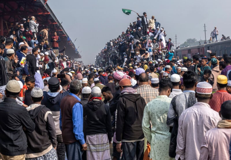 A crowded train station with people covering the train's roof and platform. Many wear traditional attire and hats. The scene is bustling, with a clear sky in the background.