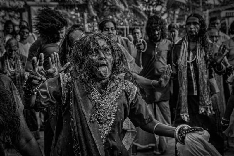 A black-and-white image shows a woman in elaborate traditional attire with messy hair, energetically dancing among a group of people. Her expression is intense, with her tongue out. The crowd behind her observes and participates in the procession.