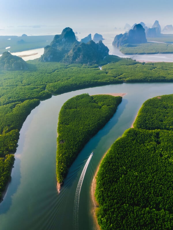 Aerial view of a boat traveling on a winding river through lush green mangrove forests. The scene is framed by distant rocky mountains under a clear blue sky, creating a serene and picturesque landscape.