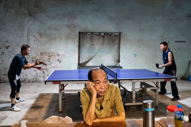 An older man in a yellow shirt sits with his head resting on his hand, looking thoughtful, while two people play table tennis in the background in an old room with bare walls. A table with a flask and cup is in the foreground.