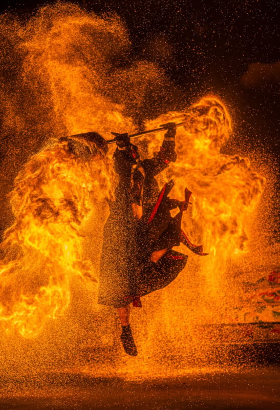 Performer in dark clothing executing a dynamic, fiery dance with flames swirling around. The intense motion creates a dramatic scene with sparks illuminating the background, highlighting the energy and movement.