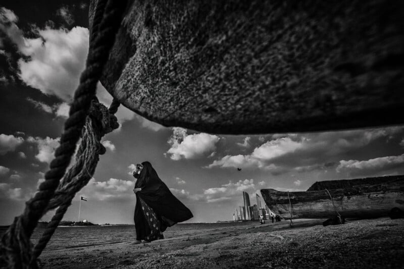 A person in a dark robe walks along a sandy shore, framed by an old boat in the foreground. In the background, modern skyscrapers rise under a cloudy sky. A flag flies on a pole near the horizon. The scene is in black and white.