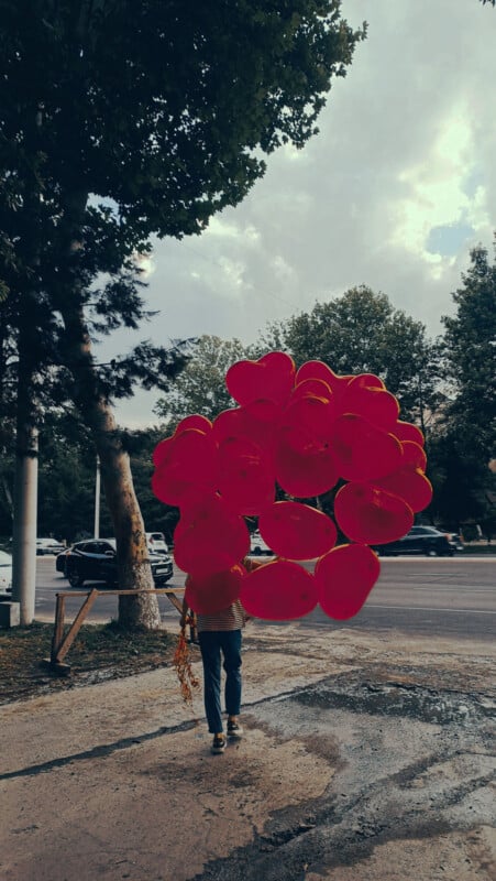 A person walking on a sidewalk carrying a large bunch of red heart-shaped balloons. Trees and parked cars line the street in the background under a cloudy sky.