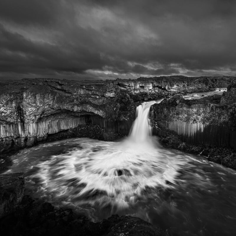 Dramatic black and white photo of a powerful waterfall cascading over rugged cliffs into a swirling pool below. Dark, brooding clouds loom overhead, creating a striking, moody atmosphere.