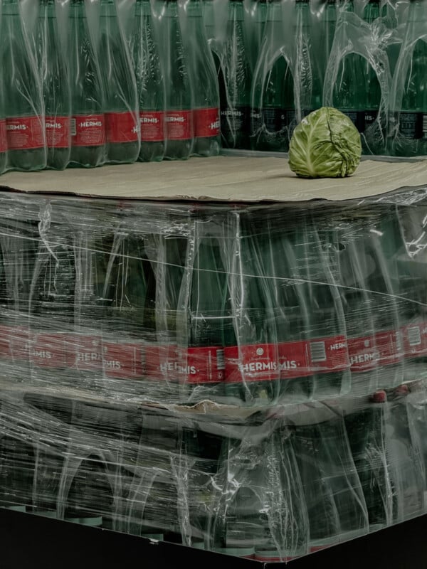 A large stack of wrapped plastic bottles with red labels sits on a pallet. On top of the pallet, a single cabbage is placed, standing out against the uniformity of the bottles.