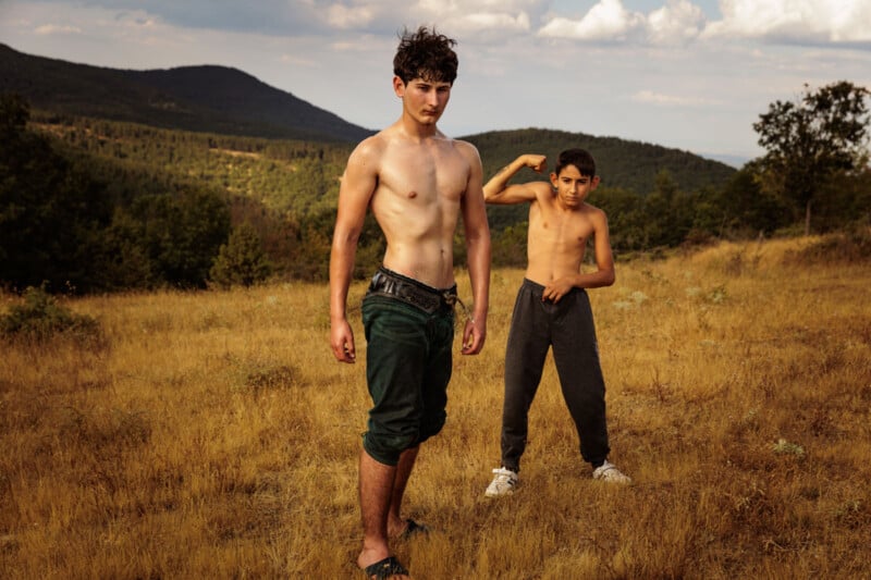 Two shirtless boys stand on a grassy field. The older boy looks ahead seriously, while the younger one flexes his arm muscle. In the background, there are trees and hills under a partly cloudy sky.