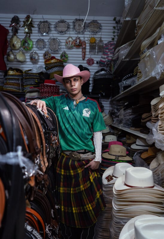 A person wearing a pink cowboy hat, green soccer jersey, and plaid skirt stands in a hat store. They lean against a shelf filled with belts and hats, surrounded by various styles of headwear hanging on the walls.