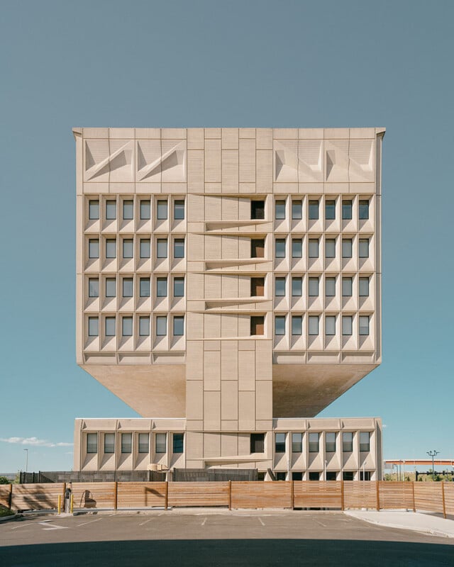 A modern, symmetrical building with a geometric design, featuring square windows and an elevated base. The structure stands against a clear blue sky, with an empty parking area in the foreground.