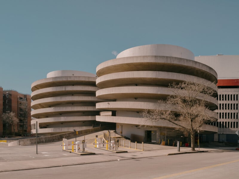 A brutalist-style multistory parking garage with two circular ramp towers. The structure is made of concrete and appears weathered. Yellow barriers and faded lines mark the parking areas. A bare tree stands near the entrance under a clear blue sky.