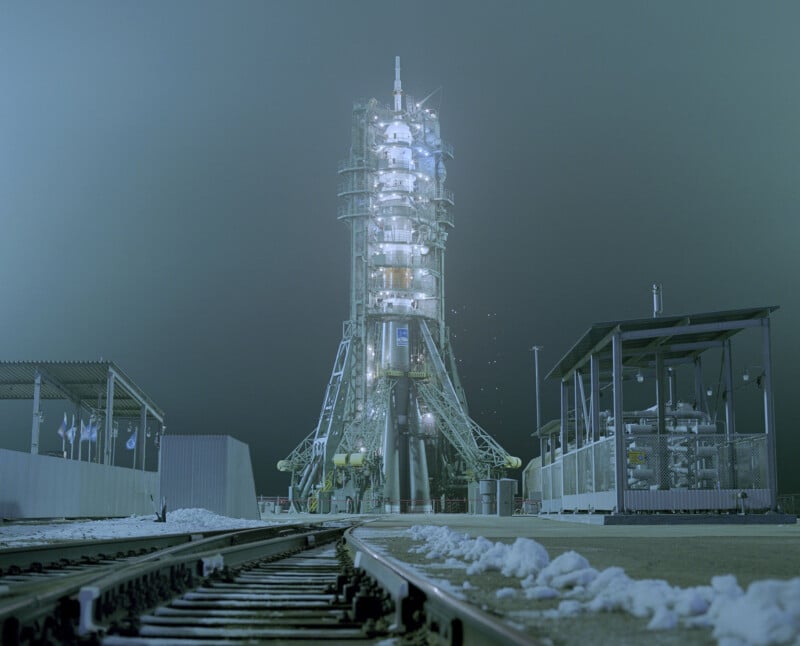 A tall rocket stands on a launch pad at night, illuminated by bright lights. The surrounding area is dusted with snow, and a set of railroad tracks leads up to the pad. Structures and equipment are visible nearby under a foggy sky.