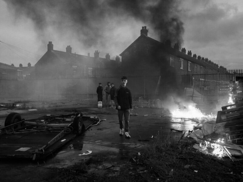 A black and white photo shows a young person walking away from large plumes of smoke and small fires near a row of houses. Nearby, a few other people stand amidst scattered debris and an overturned trailer on a wet street.