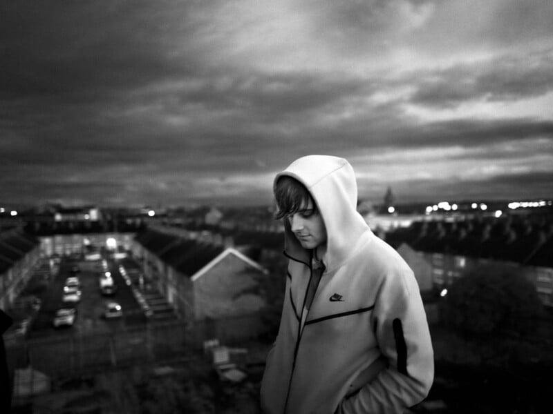 A person in a hooded jacket stands on a rooftop overlooking a blurred urban landscape with rows of houses and parked cars. The sky is overcast, and the setting is in black and white.