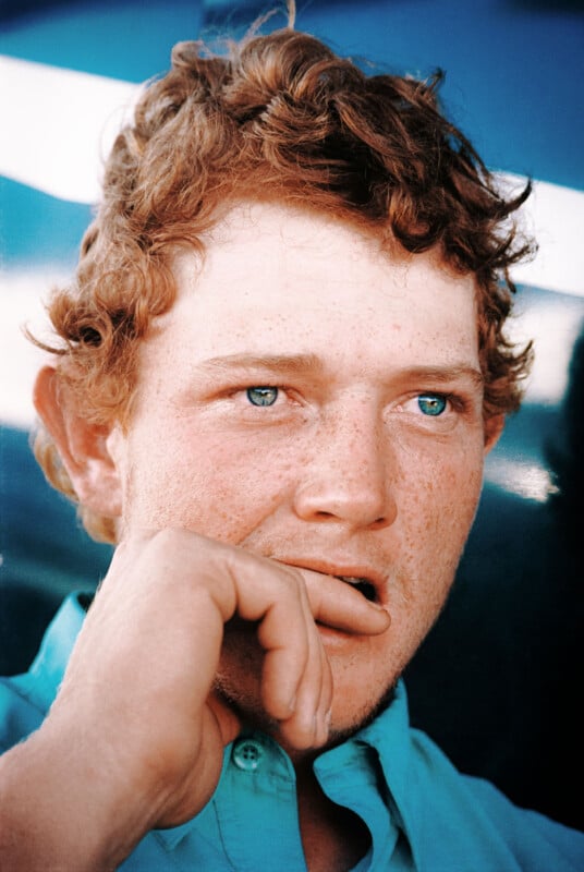 A person with curly red hair and blue eyes is staring off to the side, their hand partially covering their mouth. They are wearing a light blue shirt. The background is blurred with shades of blue and white.