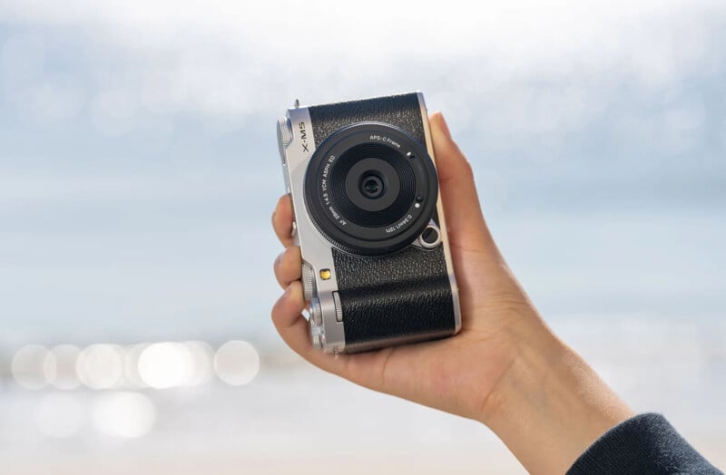 A hand holding a compact black and silver camera against a blurred beach background. The camera has a textured grip and a lens with a silver rim, focusing on the central lens area. Bright sunlight creates a soft bokeh effect.