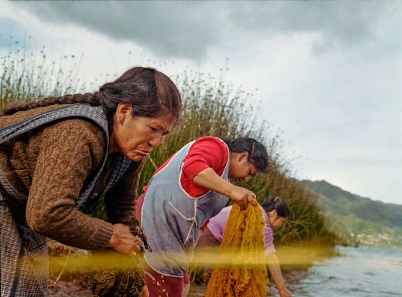 Three women stand by a lakeshore, washing clothes. Two are bent over, scrubbing with determination. Tall grasses surround them, with mountains visible in the distance under a cloudy sky.