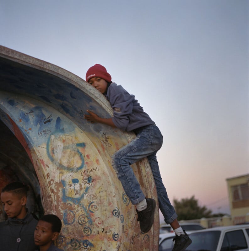 A child is perched on a graffiti-covered, arched concrete structure, wearing a red beanie and denim jeans. Two other children stand nearby, looking away. The sky is clear and houses are visible in the background.