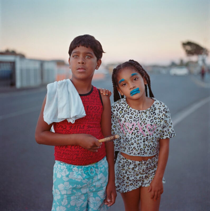 A young boy holds a stick, with a cloth draped over his shoulder, wearing a red and black top and blue floral shorts. Beside him, a girl wears a leopard print outfit with blue lipstick. They stand on a street with a soft evening sky in the background.