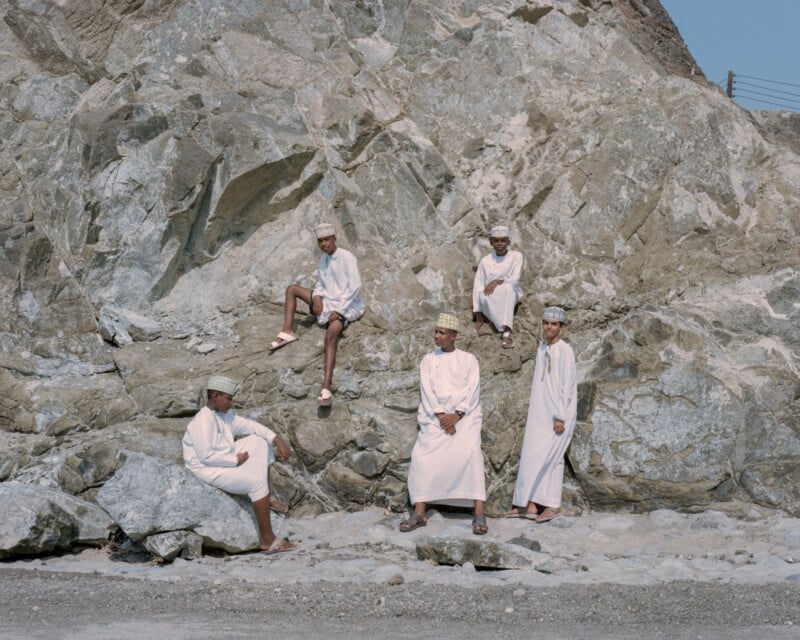 Five people in traditional attire sit and stand on large rock formations. The scene depicts a relaxed, natural setting with rugged, textured rocks as the backdrop, and a clear sky above.