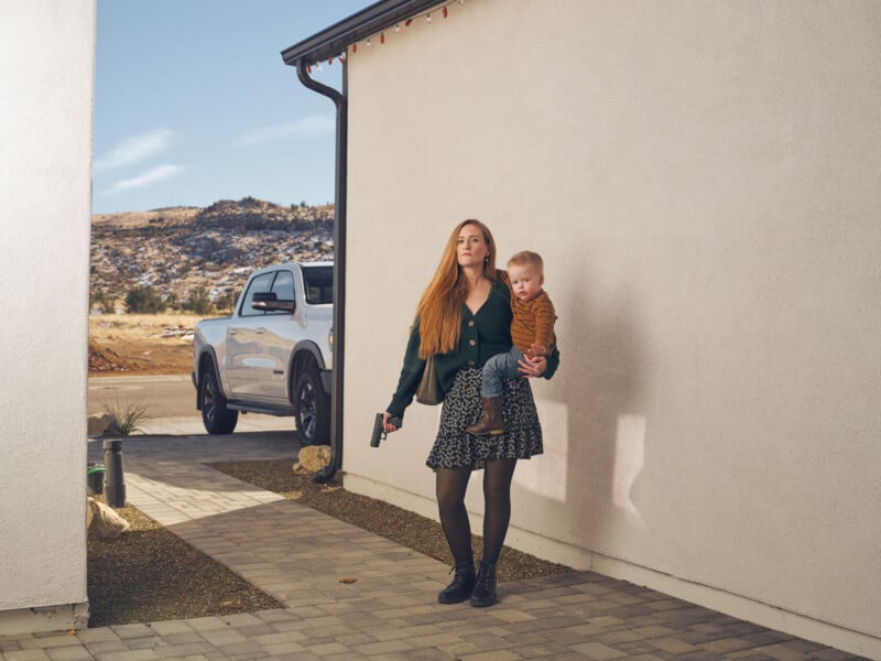 A woman with long hair, wearing a patterned dress and boots, holds a toddler in one arm and a handgun in the other. They stand outside a house next to a white vehicle, with a desert landscape in the background.