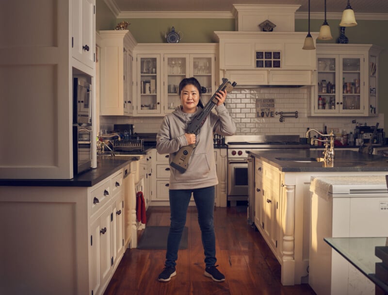 A person stands in a kitchen, holding a Fortnite-themed Nerf gun. The kitchen features white cabinets, a tiled backsplash, and hanging lights. They are wearing a gray hoodie and jeans, standing on a wooden floor.