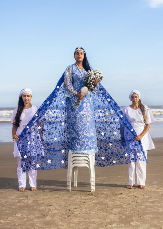 A woman in a blue, intricately patterned dress stands on a chair at the beach, holding flowers. Her dress extends with a lace-like cape held by two people dressed in white, one on each side. The ocean and sky are visible in the background.