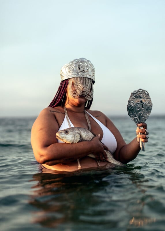 A person wearing a white bikini and ornate silver headdress stands in the ocean. They hold a fish in one hand and an intricate silver object resembling a mirror in the other. The sky is clear and the water is calm.