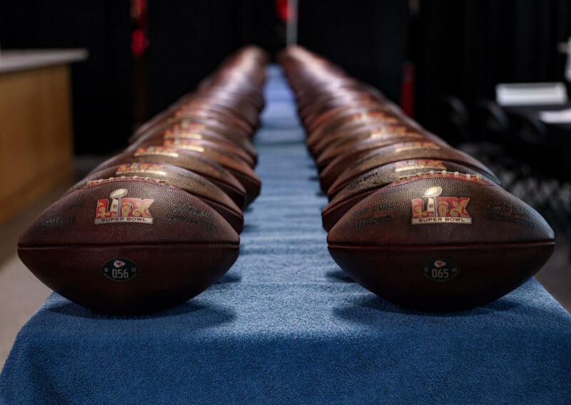 A row of Super Bowl footballs is neatly arranged on top of a blue cloth-covered table. The footballs have the Super Bowl logo and are aligned in a way that creates a striking visual perspective.