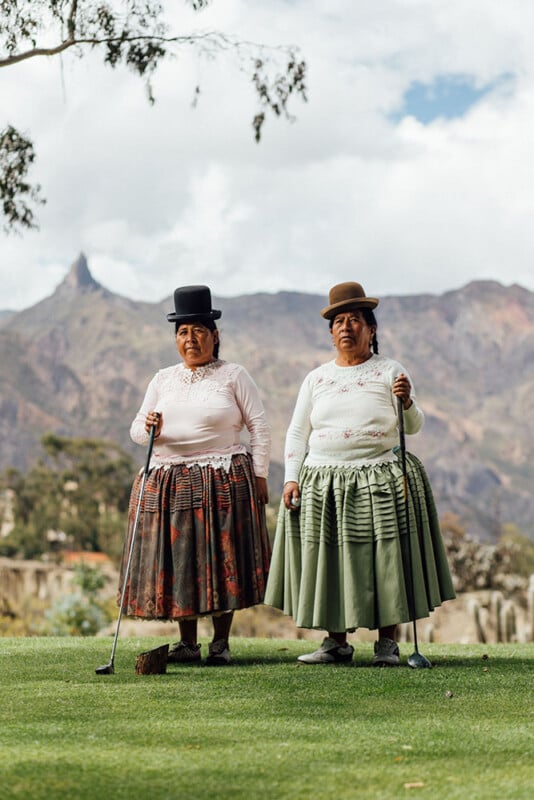 Two women in traditional attire and bowler hats stand on a golf course holding clubs. They are on a green with mountainous scenery and cloudy skies in the background.