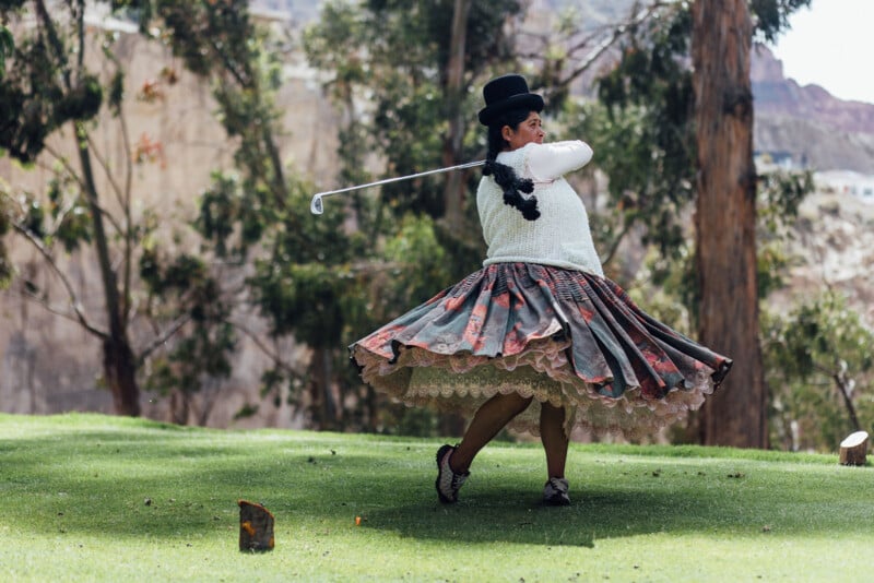 A woman in traditional attire swings a golf club on a lush green course. Her colorful, layered skirt billows around her, and she wears a bowler hat. Tall trees are visible in the background.