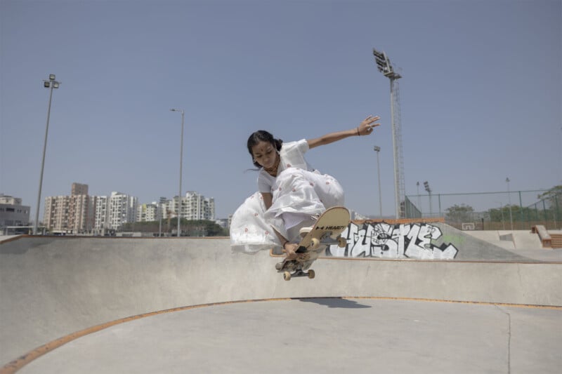 A young person in a white dress performs a skateboard trick mid-air in a skatepark, with buildings and graffiti visible in the background under a clear blue sky.