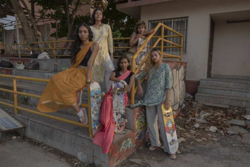 A group of five women in saris stand and sit on a set of concrete steps next to yellow railings. Each holds a skateboard. The background shows a building and some trees. The scene has a serene and empowering vibe.