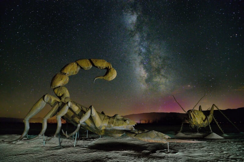 Massive metal sculptures of a scorpion and a grasshopper sit under a star-filled night sky with the Milky Way visible. The ground is sandy, and the distant horizon glows with faint, colorful lights.