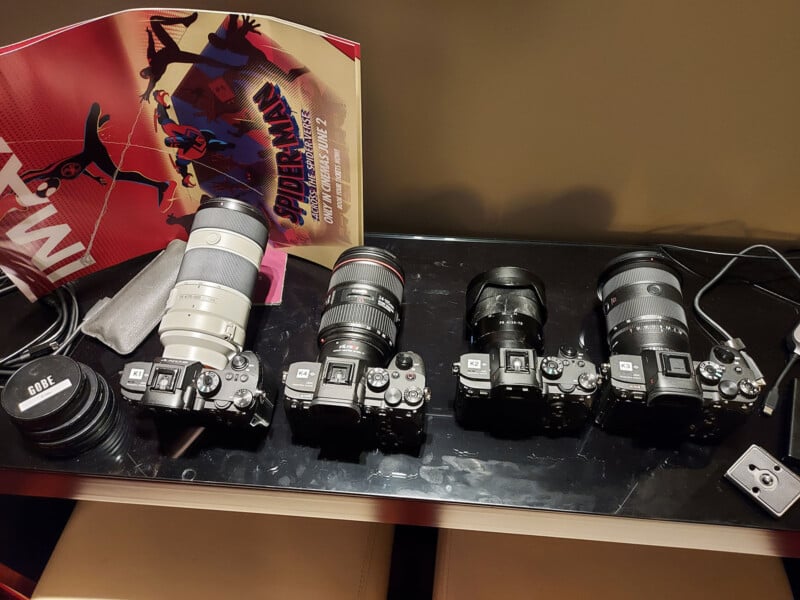 A row of four cameras with various lenses is displayed on a dark surface. Behind them, a colorful "Spider-Man: Across the Spider-Verse" poster is partially visible. Some camera accessories and cables are also present.