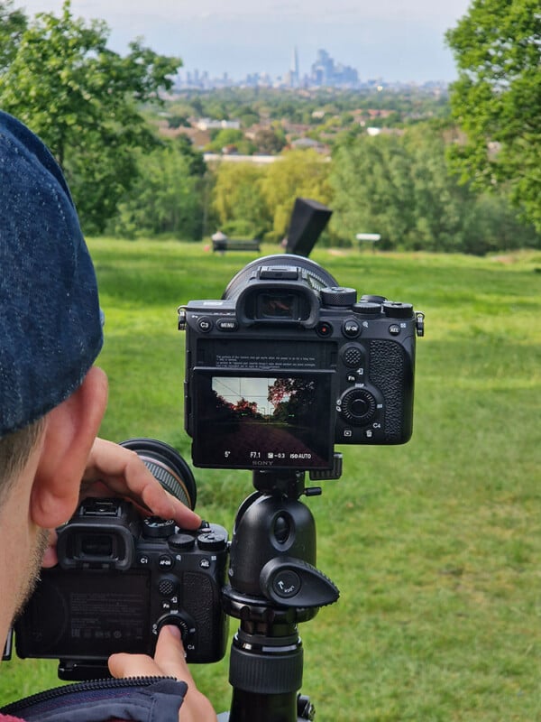 A person is taking a photo of a city skyline in the distance using a professional camera mounted on a tripod. The camera's screen displays the same city view, surrounded by green trees and grass in the foreground.