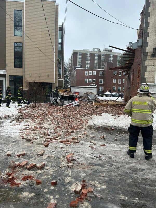 A collapsed building with debris and bricks scattered on the snowy ground. Several firefighters in uniform are present, assessing the damage. Nearby buildings are visible, and the road is icy.