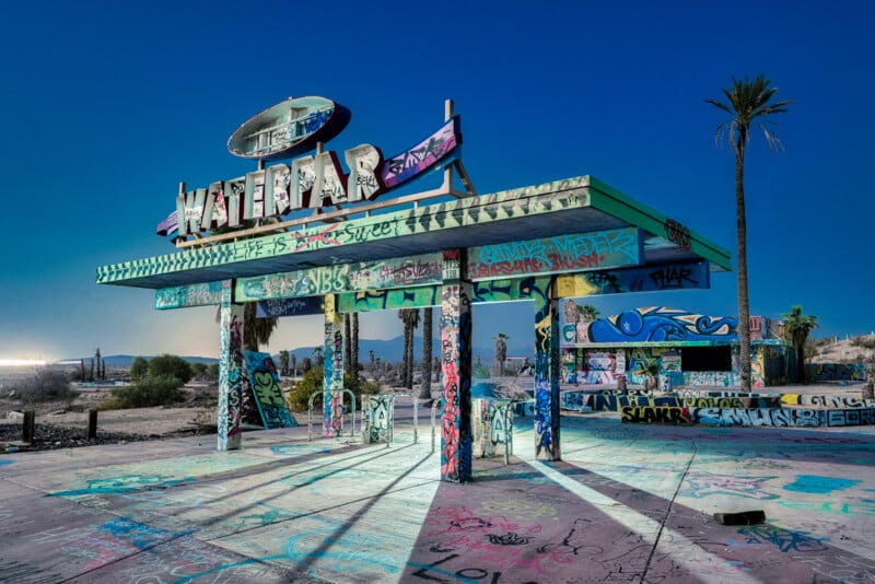 Abandoned gas station covered in colorful graffiti, with a large, weathered sign reading "Motel" at the top. The area is illuminated, surrounded by desert landscape, and a single palm tree stands nearby under a clear blue sky.