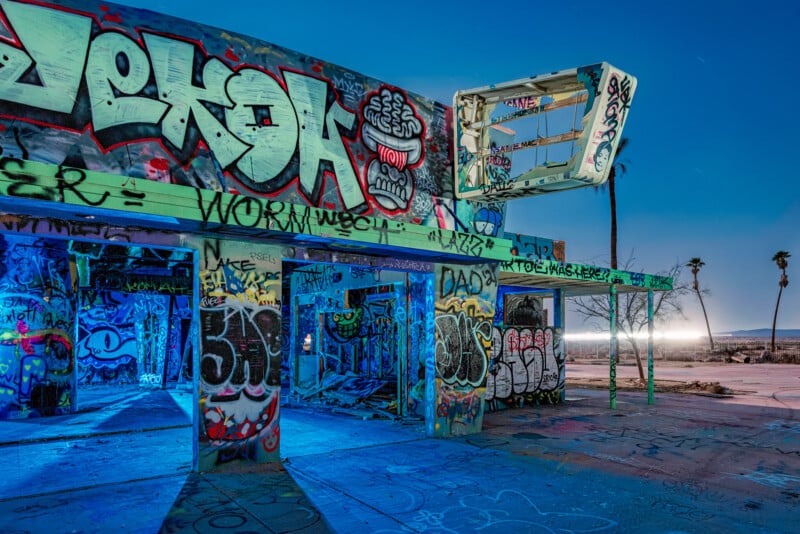 Graffiti-covered abandoned building illuminated in blue light during the evening. The structure has exposed walls and roof beams, with palm trees visible in the background under a clear sky.