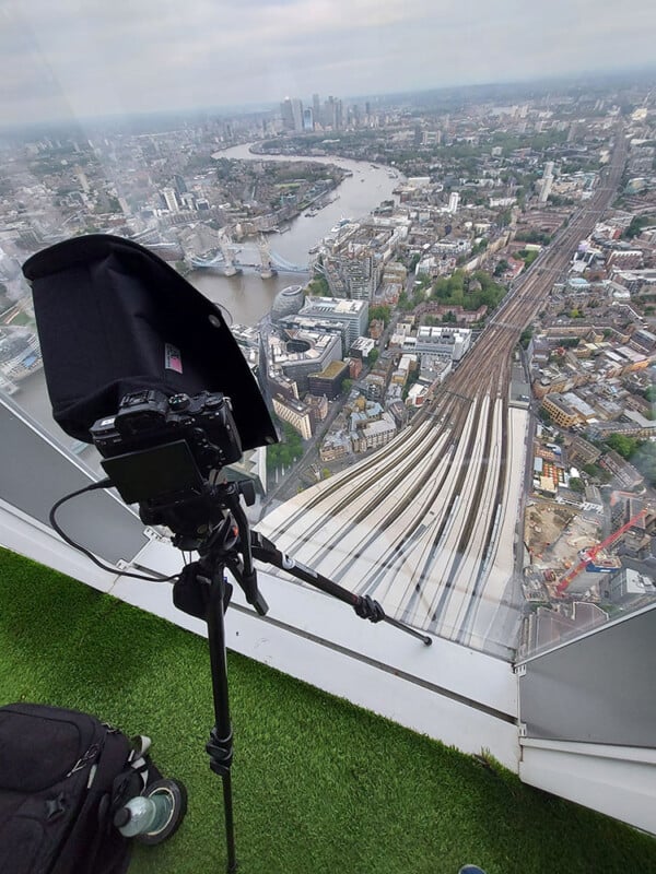 A camera on a tripod positioned near a glass wall offers a bird's eye view of a cityscape with railway tracks, the Thames River, and surrounding buildings. The vantage point includes a patch of artificial grass.