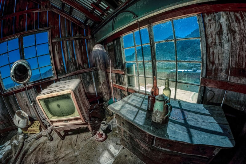 Interior of a rustic, abandoned room with vintage items. A dusty TV stands by the wooden wall, a lampshade overhead. On the wooden table, two bottles and a rusty jug sit. Sunlight streams through large windows with a mountain view.
