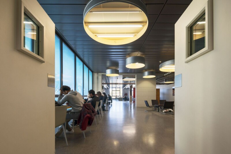 A well-lit common area with large circular lights hanging from the ceiling. People are seated at tables near floor-to-ceiling windows, engaged in study or work. Modern design elements with a corridor leading to other parts of the building.